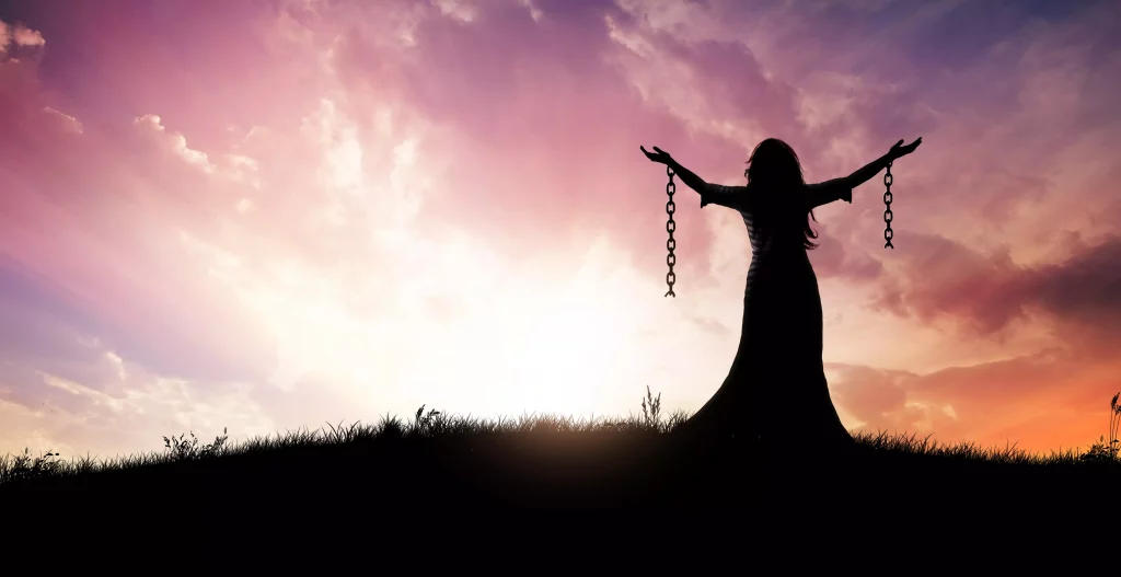 woman standing with broken chains looking into a bright sunset