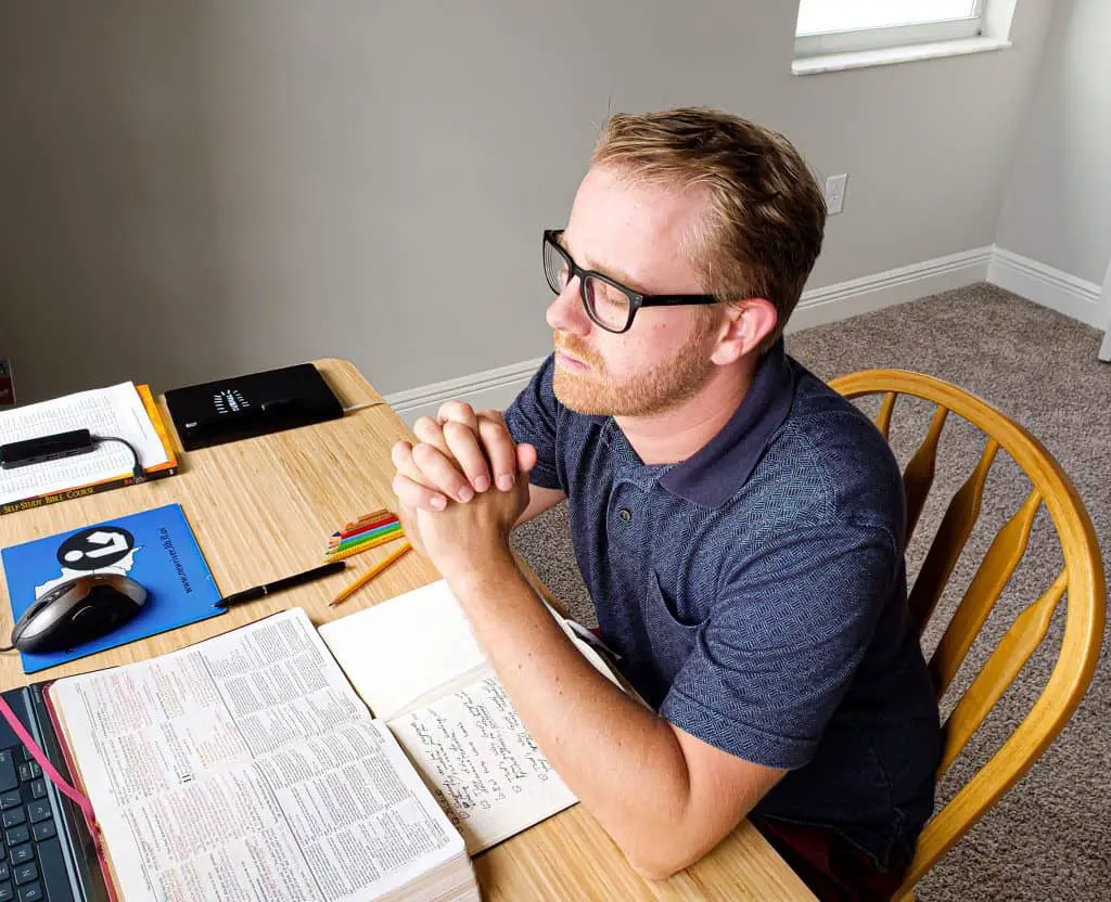 Blond man with glasses praying with folded hands sitting at an office desk with his Bible and journal opened