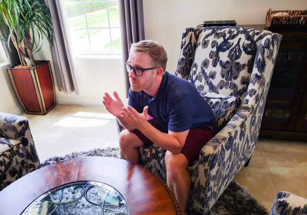 Blond man with glasses sitting in an armchair raising his hands with his elbows resting on his knees.