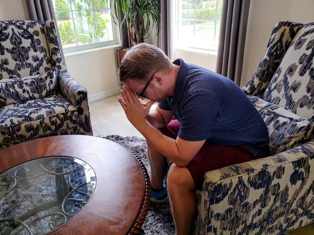 Man sitting at a round living room table in an armchair hunched over with praying hands touching his forehead.