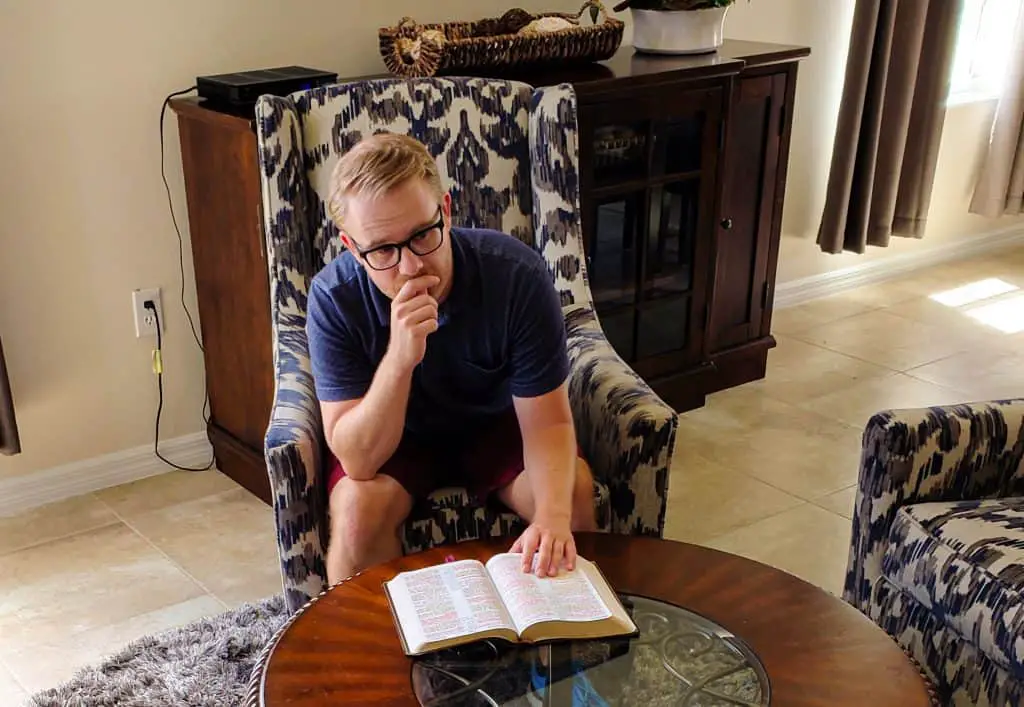 Man sitting at a living room table with an open Bible in front of him on the table. His tight hand is touching his chin in a thoughtful pose.