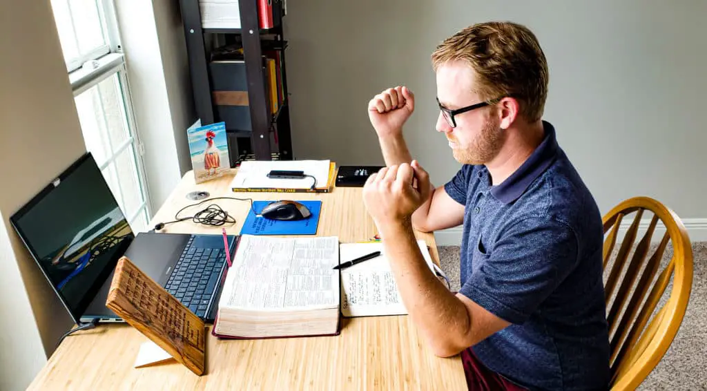 Man sitting at a desk. He rests his elbows on the desk with his fists going straight up in the air. In front of him is an open Bible, open journal, and a Laptop.