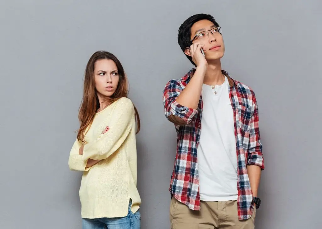 Portrait of upset angry girl watching her boyfriend talking on mobile phone isolated on the gray background