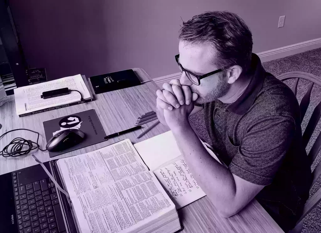 Man sitting at a desk resting his elbows on the desk. His hands are folded in front of his mouth in a prayer pose. in front of him are an open Bible and open Journal.