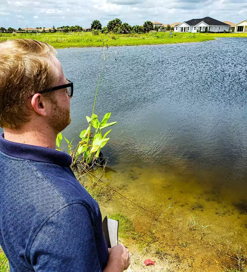 Blond mand overlooking pond with bible in his hand houses in the background