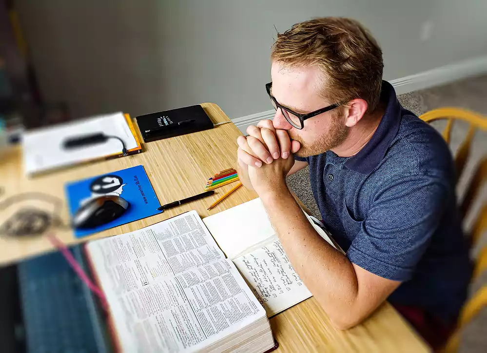 man praying at a desk resting his his chin on his folded hands, in front of him is an open Bible