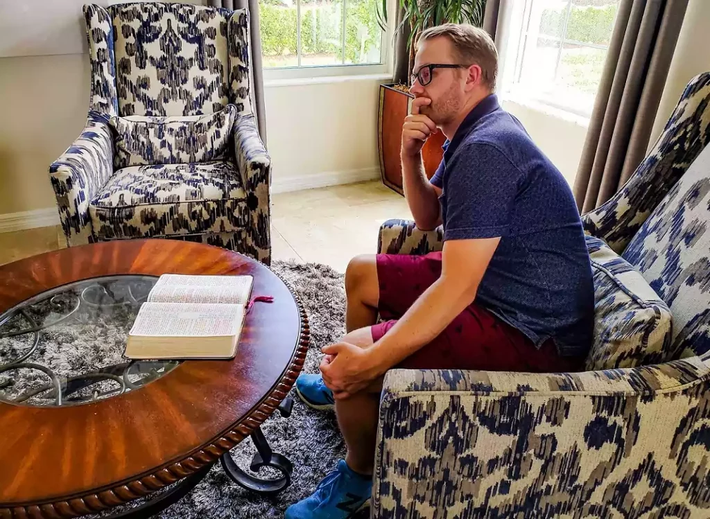 man sits in an arm chair in thinking pose with his bible opened in front of him laying on a coffee table.