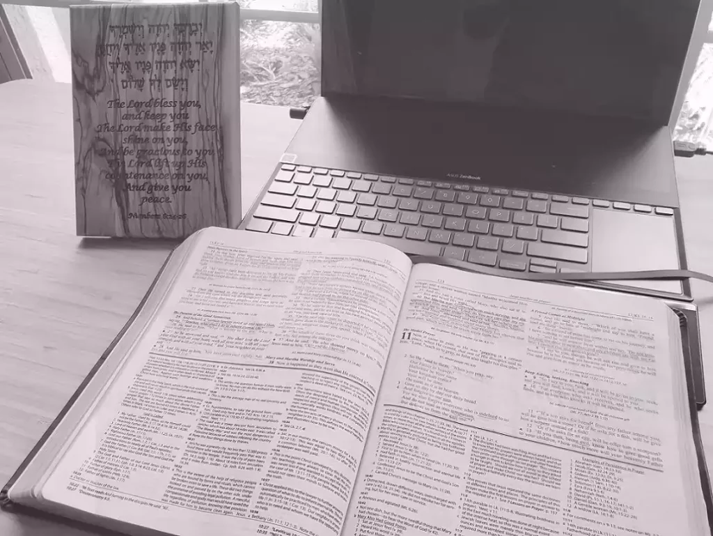 An open Bible lies in front of a laptop on a desk. Next to the laptop is a wooden plaque with Hebrew letters